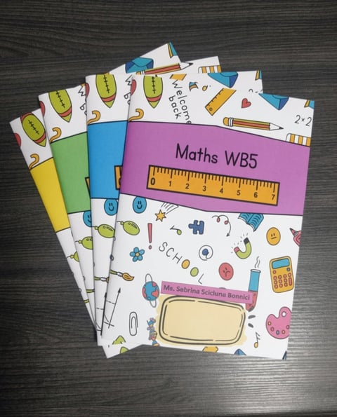 Stack of colorful school workbooks including a Maths WB5 book with ruler, labeled Ms. Sabrina Bonnici, featuring educational icons and patterns on a gray surface.