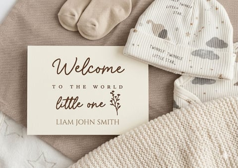 Welcome to the world card with baby items including beige socks, patterned beanie, and cream blanket on neutral fabric background